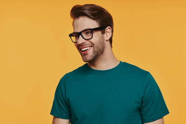 Veselý mladý muž v brýlích dívá pryč a usmívá se, zatímco stojí na žlutém pozadí — Stock fotografie