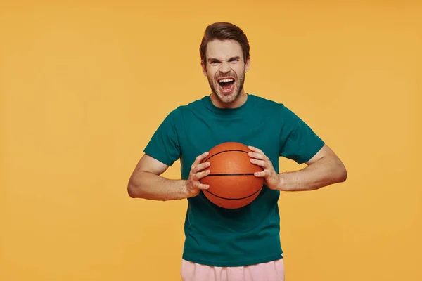 Furioso giovane uomo in possesso di palla da basket e guardando la fotocamera mentre in piedi contro sfondo giallo — Foto Stock