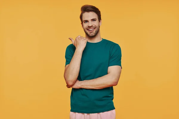 Handsome young smiling man in casual clothing looking at camera and pointing away — Stock Photo, Image