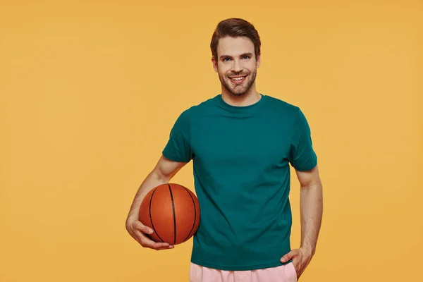 Guapo joven sosteniendo pelota de baloncesto y sonriendo mientras está de pie sobre fondo amarillo —  Fotos de Stock