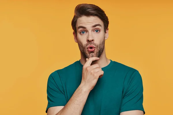 Surprised young man holding hand on chin and making a face while standing against yellow background — Stock Photo, Image