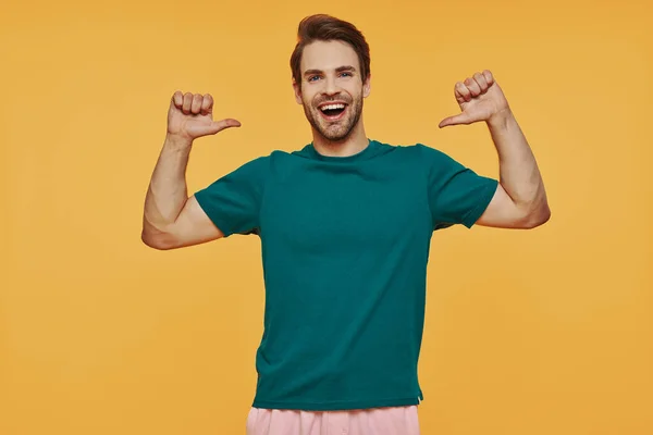 Bonito jovem sorrindo homem em roupas casuais olhando para a câmera e apontando-se — Fotografia de Stock