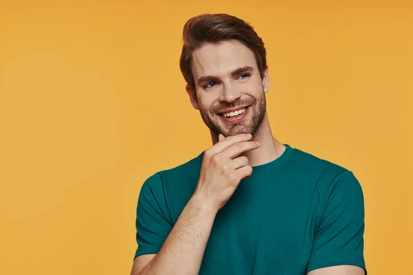 Thoughtful young man holding hand on chin and smiling while standing against yellow background — Stock Photo, Image