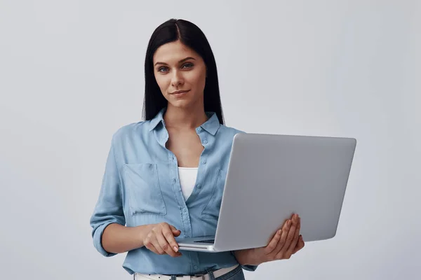 Atrakcyjna młoda kobieta korzystająca z laptopa i patrząca w kamerę stojąc na szarym tle — Zdjęcie stockowe