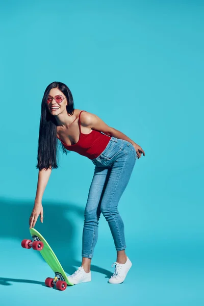 Full length of attractive young woman looking at camera and smiling while skateboarding against blue background — 图库照片