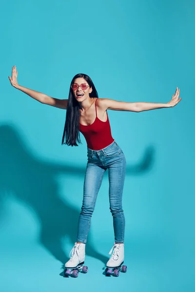 Full length of attractive young woman looking at camera and smiling while skating against blue background — 图库照片