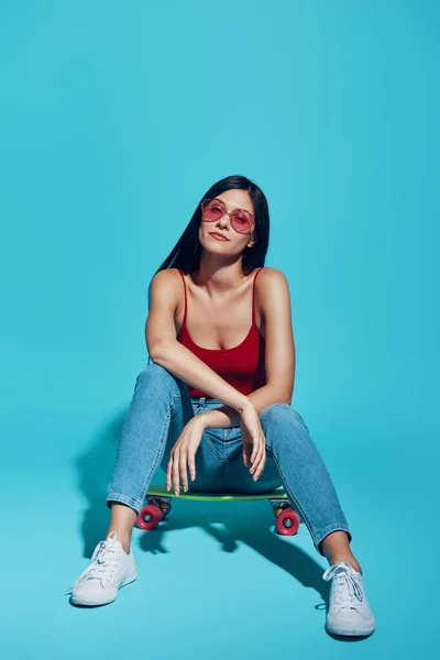 Attractive young woman looking at camera while sitting on skateboard against blue background — 图库照片