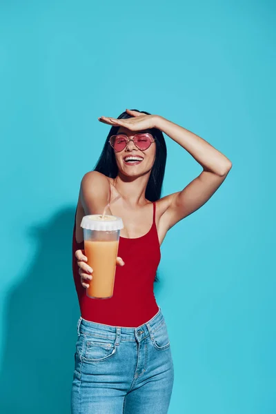 Приваблива молода жінка посміхається і дає вам коктейль, стоячи на синьому фоні — стокове фото