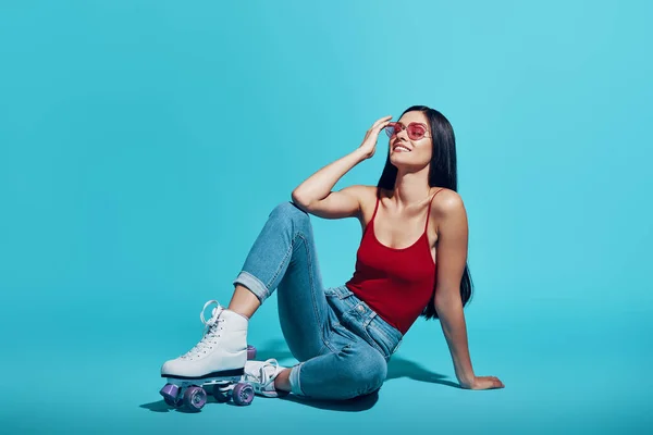 Attractive young woman wearing roller skates and smiling while sitting against blue background — 图库照片