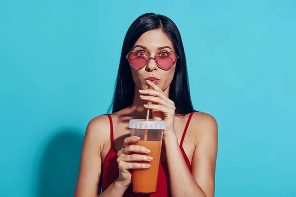 Attrayant jeune femme boire un cocktail et regarder la caméra tout en se tenant debout sur fond bleu — Photo