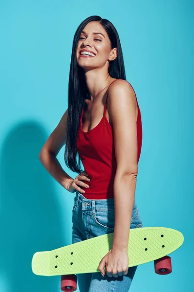 青を背景に立ちながらスケートボードと笑顔を運ぶ魅力的な若い女性 — ストック写真