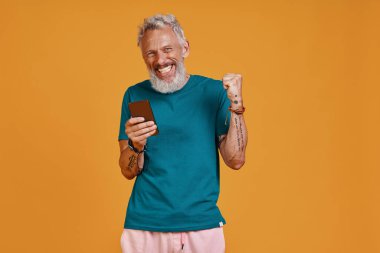 Mutlu kıdemli adam akıllı telefon kullanıyor ve turuncu arka planda dururken gülümsüyor.