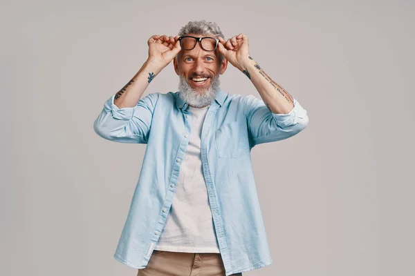Bello uomo anziano regolazione occhiali e sorridente mentre in piedi contro sfondo grigio — Foto Stock