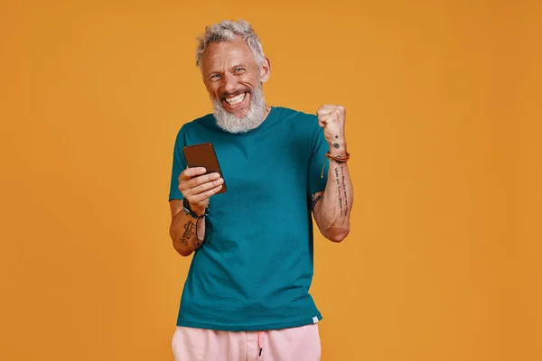 Happy senior man using smart phone and smiling while standing against orange background — Stock Photo, Image