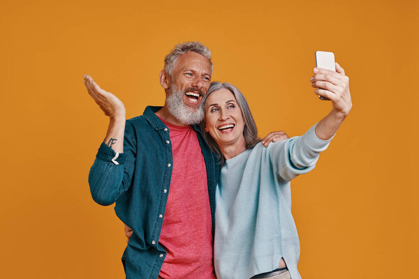 Happy senior couple using smart to take a selfie while standing together against orange background