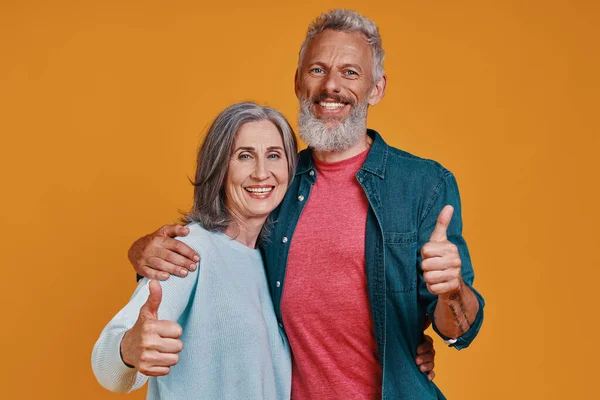 Beautiful senior couple looking at camera and keeping thumbs up while standing together against orange background — Stock Photo, Image