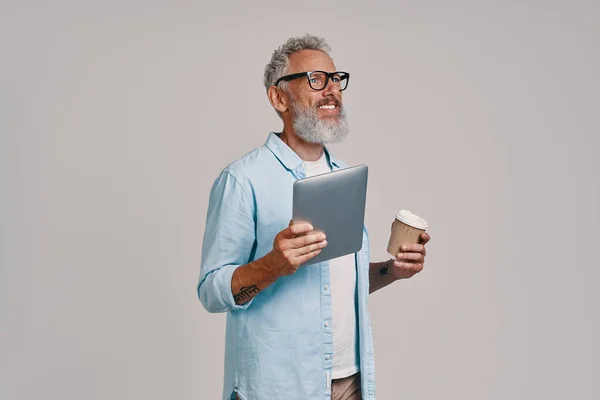 Šťastný senior muž drží digitální tablet a šálek kávy, zatímco stojí na šedém pozadí — Stock fotografie