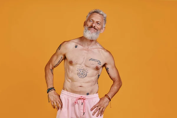 Мужчина без рубашки с татуировкой смотрит в сторону и улыбается, стоя на оранжевом фоне — стоковое фото
