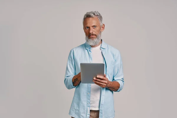 Уверенный пожилой человек с помощью цифрового планшета, стоя на сером фоне — стоковое фото