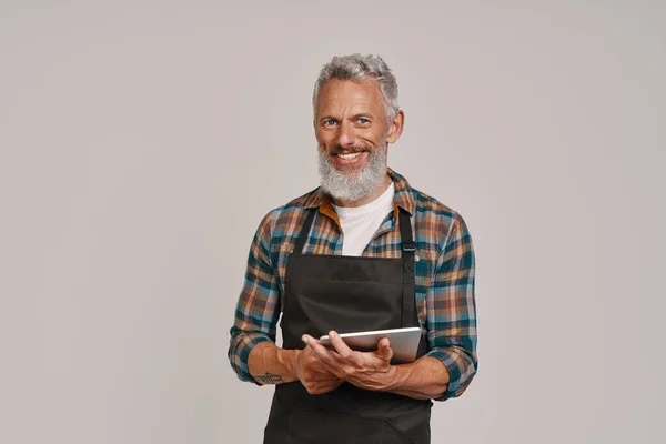 Homme âgé en tablier souriant et en utilisant une tablette numérique tout en se tenant debout sur fond gris — Photo