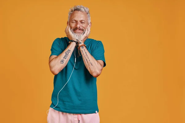 Happy senior man listening music and smiling while standing against orange background — Stock Photo, Image