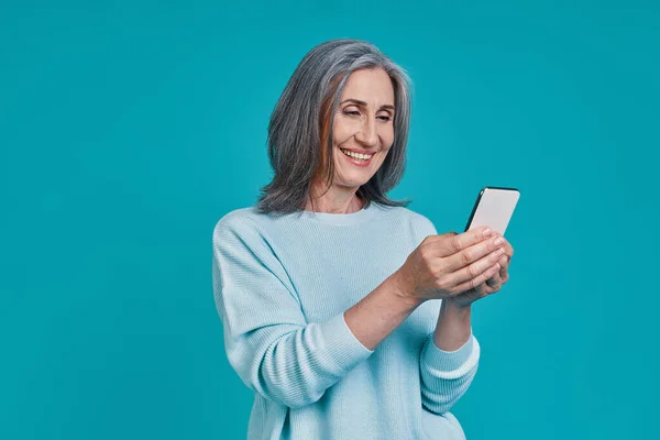 Madura hermosa mujer usando el teléfono inteligente y sonriendo, mientras que de pie sobre fondo azul — Foto de Stock