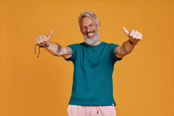 Feliz homem sênior mostrando seus polegares para cima, enquanto de pé contra o fundo laranja — Fotografia de Stock