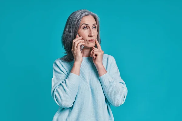 Mulher bonita madura falando no telefone inteligente enquanto está de pé contra o fundo azul — Fotografia de Stock