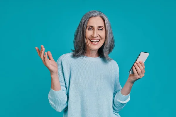 Mature beautiful woman using smart phone and smiling while standing against blue background — Stock Photo, Image