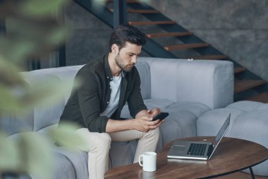 Akıllı telefon kullanan düşünceli genç adam kanepede otururken ona bakıyor.