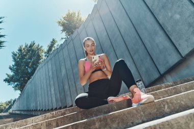 Spor elbiseli genç bir kadının açık havada oturması ve akıllı telefonlara bakması.