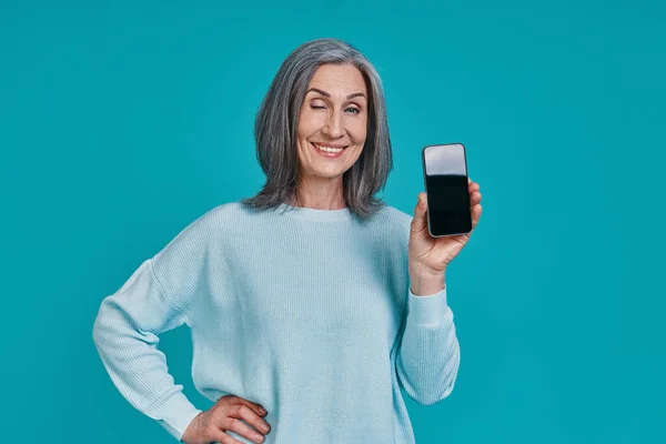 Mulher bonita madura mostrando seu telefone inteligente e sorrindo enquanto está de pé contra o fundo azul — Fotografia de Stock
