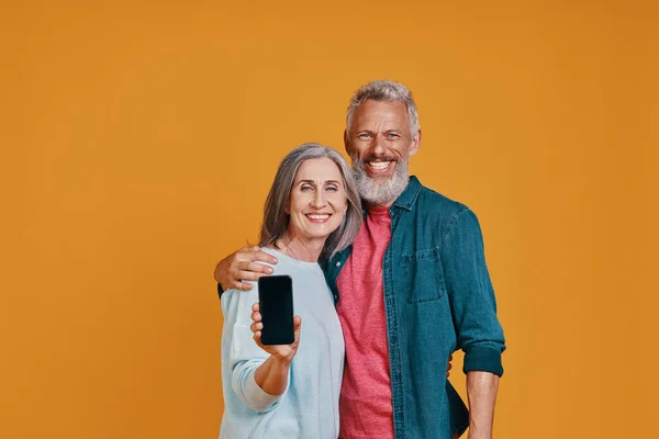 Feliz pareja de ancianos sonriendo y mostrando el teléfono inteligente mientras están de pie juntos contra el fondo naranja — Foto de Stock