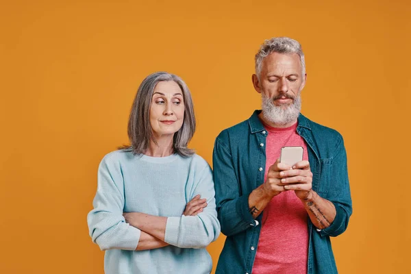 Vacker senior kvinna kikar på sin man medan han använder smart telefon mot orange bakgrund — Stockfoto