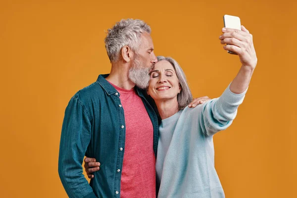 Beautiful senior couple using smart to take a selfie while standing together against orange background — Stock Photo, Image