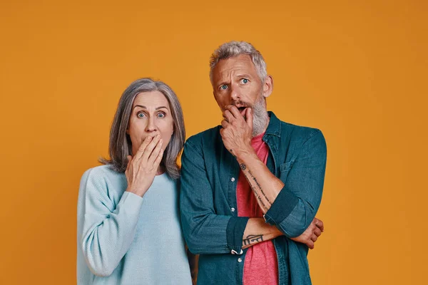 Överraskad senior par täcker munnen med handen när du står tillsammans mot orange bakgrund — Stockfoto