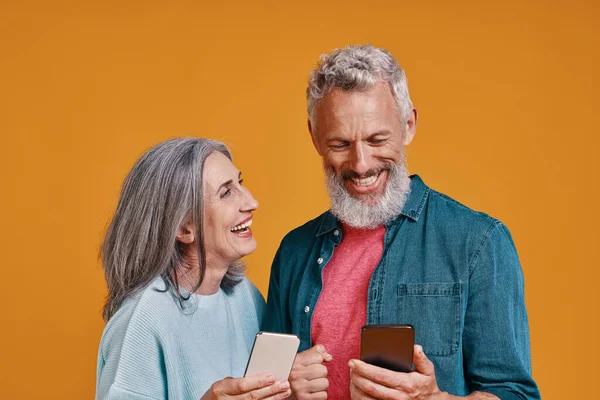 Casal sênior feliz sorrindo e usando telefones inteligentes enquanto estão juntos contra o fundo laranja — Fotografia de Stock