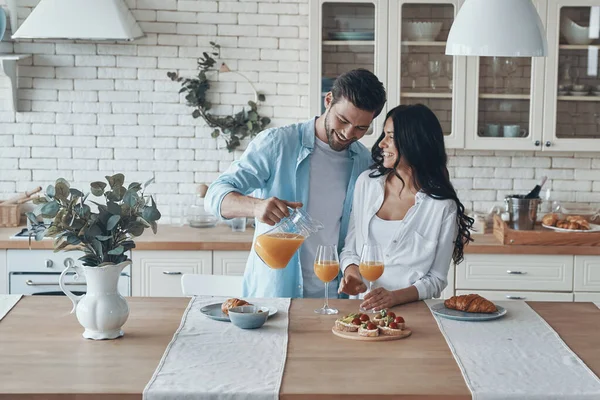 Όμορφος νεαρός ρίχνει χυμό πορτοκάλι, ενώ ετοιμάζει πρωινό μαζί με την κοπέλα του — Φωτογραφία Αρχείου