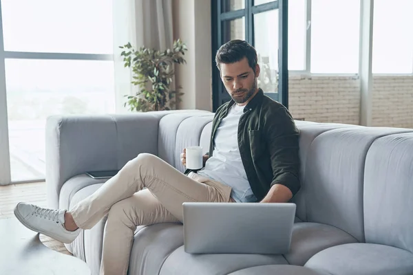 Hombre joven guapo usando el ordenador portátil y beber café mientras está sentado en el sofá en casa — Foto de Stock