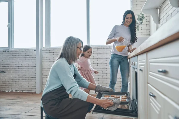 Feliz mujer mayor sacando galletas del horno y sonriendo mientras pasa tiempo con la familia —  Fotos de Stock