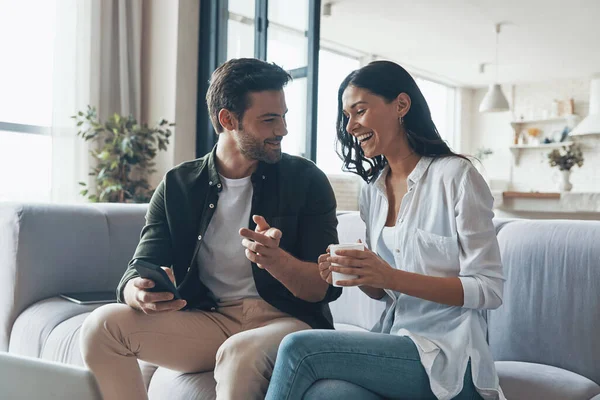 Stilig ung visar sin flickvän smart telefon och ler medan du sitter på soffan hemma — Stockfoto