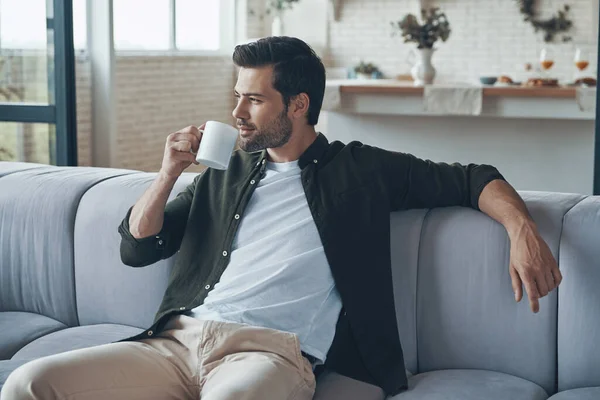 Задумчивый молодой человек пьет кофе и смотрит в сторону, сидя на диване — стоковое фото