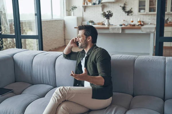 Bonito jovem falando no telefone inteligente e sorrindo enquanto sentado no sofá — Fotografia de Stock