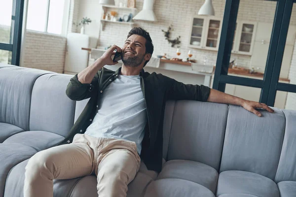 Hombre joven guapo hablando en el teléfono inteligente y sonriendo mientras está sentado en el sofá —  Fotos de Stock