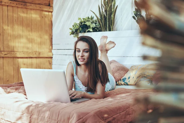 Όμορφη νεαρή χαμογελαστή γυναίκα χρησιμοποιώντας φορητό υπολογιστή, ενώ βρίσκεται στο κρεβάτι — Φωτογραφία Αρχείου