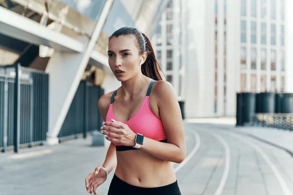 Hermosa mujer joven en ropa deportiva con auriculares y reloj inteligente mientras corre al aire libre — Foto de Stock