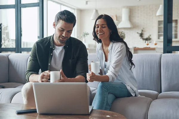 Bela jovem casal bebendo café e olhando no laptop enquanto sentado no sofá em casa — Fotografia de Stock