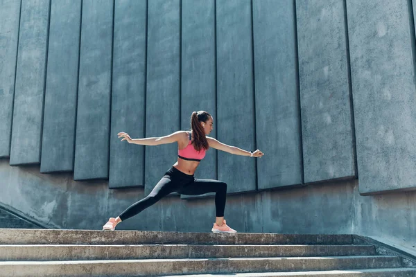 Comprimento total de jovem mulher confiante em roupas esportivas fazendo exercícios de alongamento ao ar livre — Fotografia de Stock