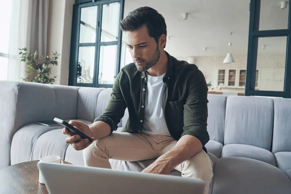 Seriózní mladý muž pomocí chytrého telefonu a notebooku, zatímco sedí na pohovce — Stock fotografie