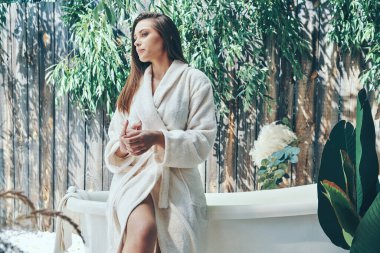 Güzel genç bir kadın, banyo küvetine yaslanırken el kremini nemlendiriyor.